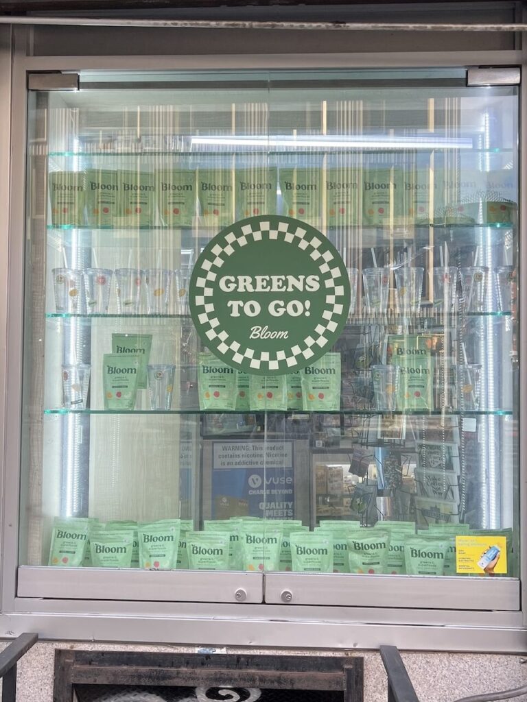 Bloom Nutrition Greens stick packs and branded cups arranged inside the storefront cooler.
